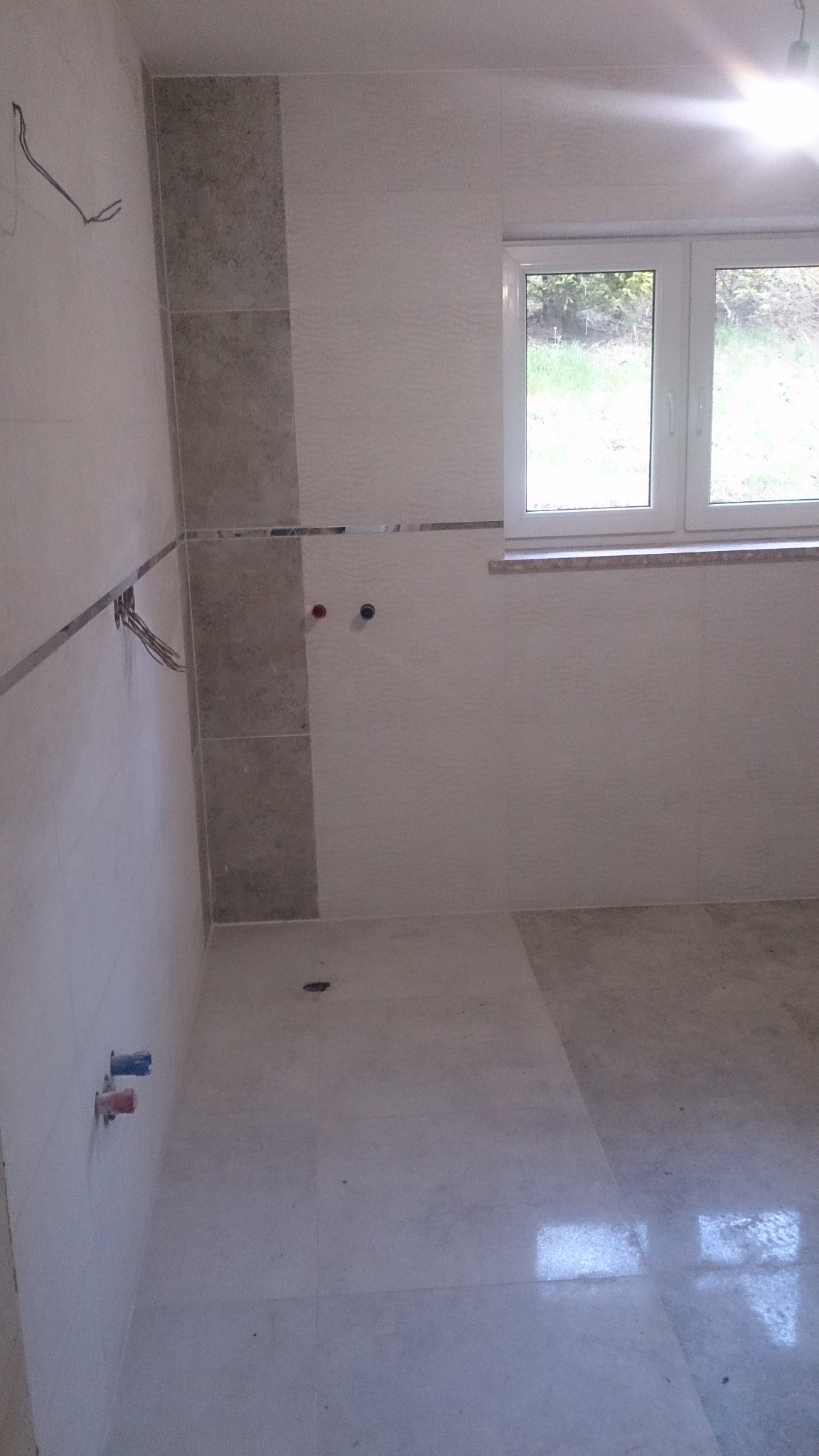 Wnętrze łazienki w trakcie wykańczania: położone jasne płytki na podłodze i ścianach, widoczne wystające kable i rury, okno z widokiem na zieleń na zewnątrz.