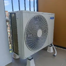 Biała jednostka zewnętrzna klimatyzacji marki AUX, zamontowana na balkonie z widokiem na niebieskie niebo, wsparta na specjalnych, białych podkładkach antywibracyjnych.