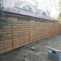 Nowo postawione, drewniane ogrodzenie panelowe z poziomych desek, widoczne na tle domu z cegły i drzew. Na ziemi leżą ślady po wycince drzew i narzędzia.