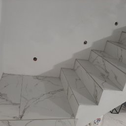 Budpat - Wykończone schody wewnętrzne z białymi stopniami i podstopnicami pokrytymi płytkami imitującymi marmur, widoczne otwory na oświetlenie w ścianie.