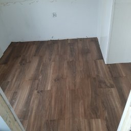 Budpat - Ułożona podłoga z płytek imitujących drewno w małym pomieszczeniu, widoczne ślady po remoncie na ścianach i gniazdko elektryczne.