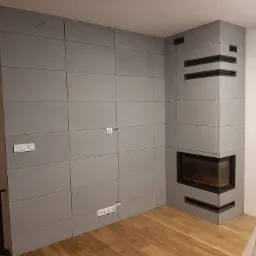 Ściana wyłożona szarymi, betonowymi płytami z widocznymi gniazdkami elektrycznymi i wbudowanym kominkiem z czarną obudową, drewniana podłoga.