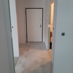 Budpat - Wykończony korytarz z ułożoną jasną, marmurkową podłogą z płytek, białe ściany i białe drzwi wejściowe z czarną ramą, widok z perspektywy niskiego kąta.