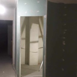 Budpat - Wnętrze w trakcie prac wykończeniowych: ściany z płyt gipsowo-kartonowych, widoczne łączenia i szpachlowanie, surowy betonowy sufit, podłoga z resztkami materiałów budowlanych, przygotowywane...