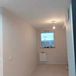 Budpat - Jasne wnętrze w trakcie prac wykończeniowych: świeżo pomalowane ściany w odcieniach bieli, nowe płytki podłogowe, grzejnik pod oknem z widokiem na budowę, surowa żarówka zwisająca z sufitu.