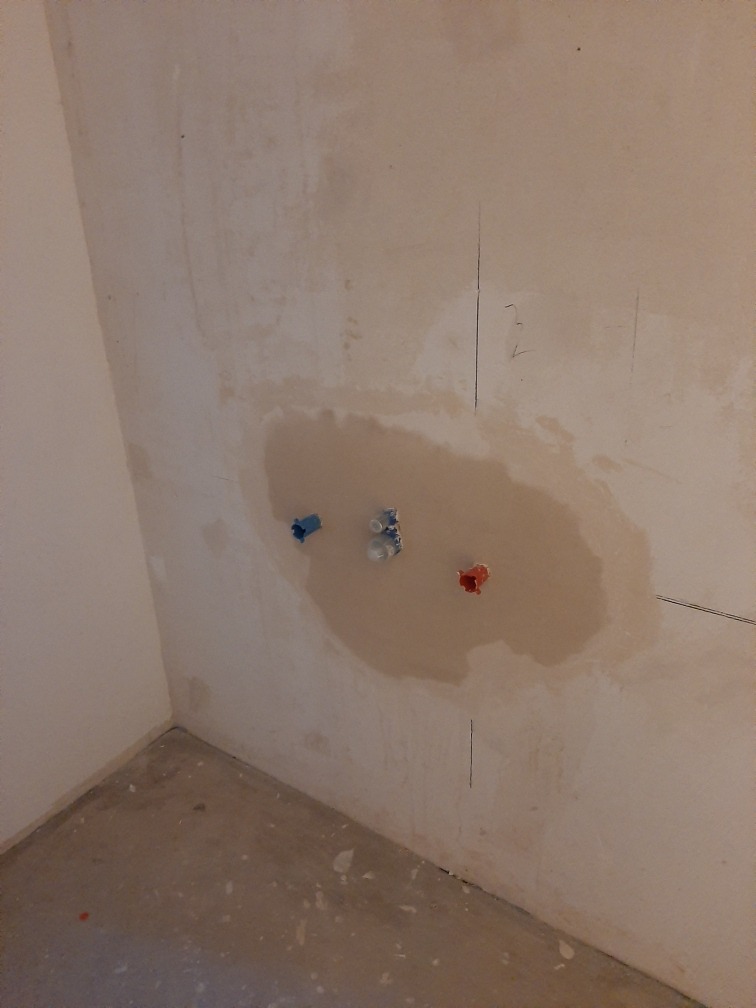 Ściana w trakcie przygotowania pod płytki w łazience, z widocznymi wyprowadzonymi przyłączami wodnymi (zimna, ciepła i odpływ) oraz śladami po łataniu ubytków w tynku.