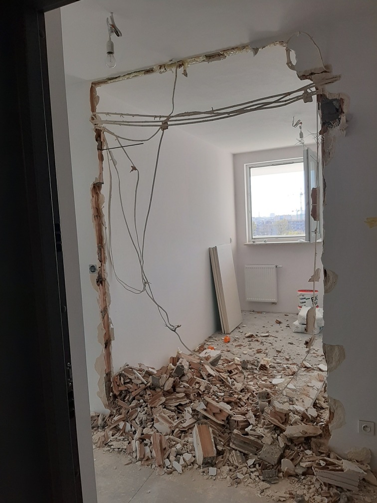 Demontaż ościeżnicy z widocznymi przewodami elektrycznymi i stertą gruzu na podłodze, ściana z widocznymi śladami po usuniętej futrynie.