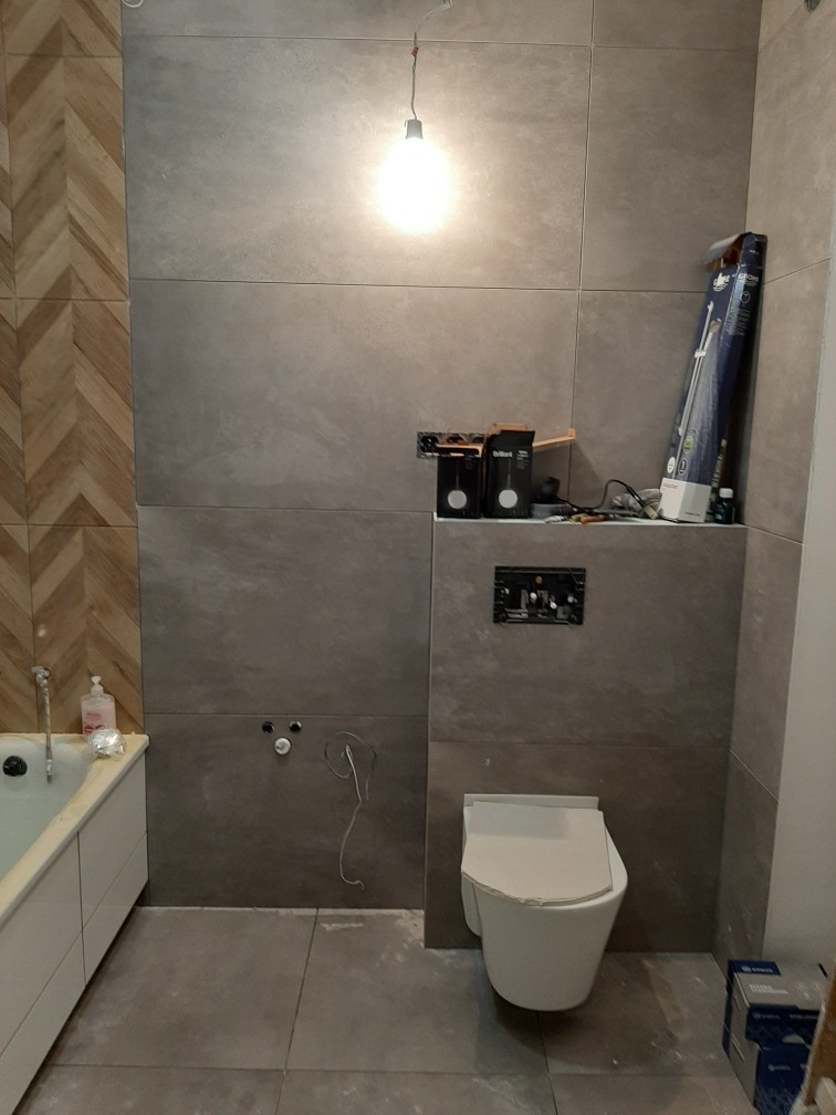 Remont łazienki w trakcie: szare płytki wielkoformatowe, częściowo ułożone, z widoczną instalacją podtynkową WC, wiszącą muszlą klozetową z zabezpieczeniem, fragmentem wanny i dekoracyjną ścianą...