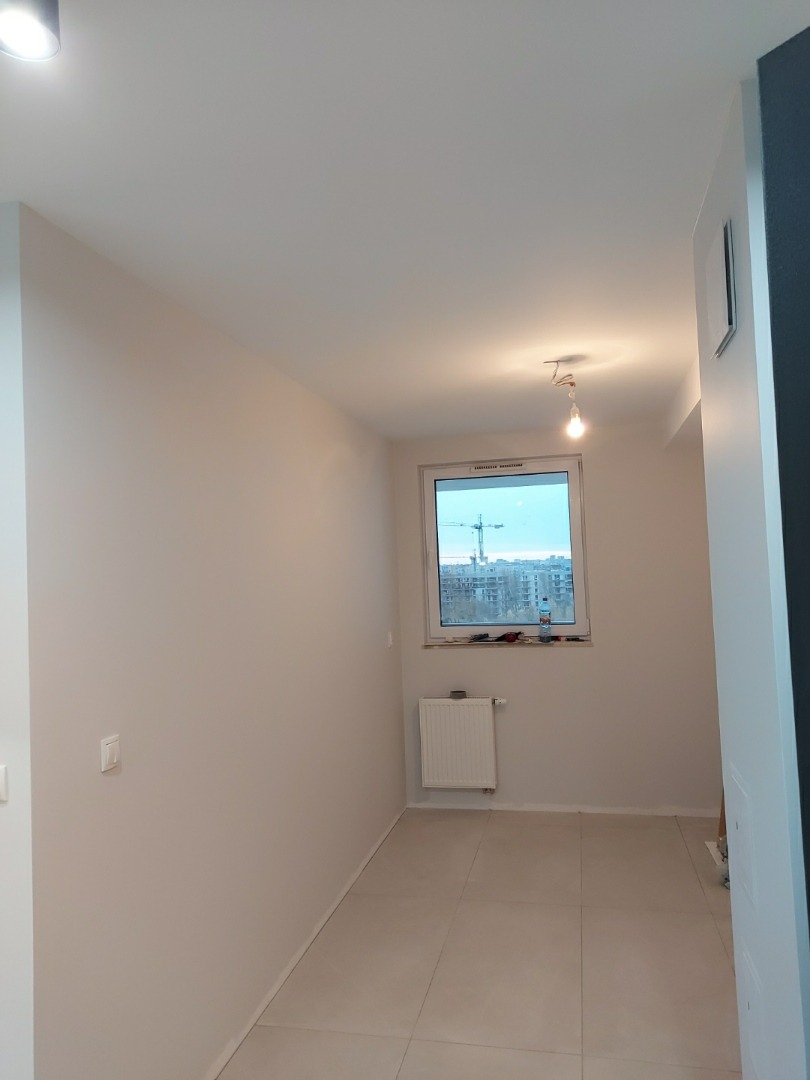 Jasne wnętrze w trakcie prac wykończeniowych: świeżo pomalowane ściany w odcieniach bieli, nowe płytki podłogowe, grzejnik pod oknem z widokiem na budowę, surowa żarówka zwisająca z sufitu.