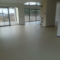 Mikrocement Podlasie - Jasna, jednolita posadzka w przestronnym, minimalistycznym wnętrzu z dużymi oknami i widokiem na zewnątrz, zabezpieczona taśmą malarską przy ścianach.