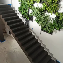 Schody pokryte szarą powłoką, z pionowym ogrodem na ścianie. Widoczne zabezpieczenia taśmą malarską. Surowy beton na podłodze. Minimalistyczny design.