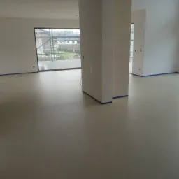 Jasnoszara, gładka podłoga w nowym, pustym pomieszczeniu z dużym oknem i kolumną. Podłoga zabezpieczona niebieską taśmą. Widok na domy przez okno.