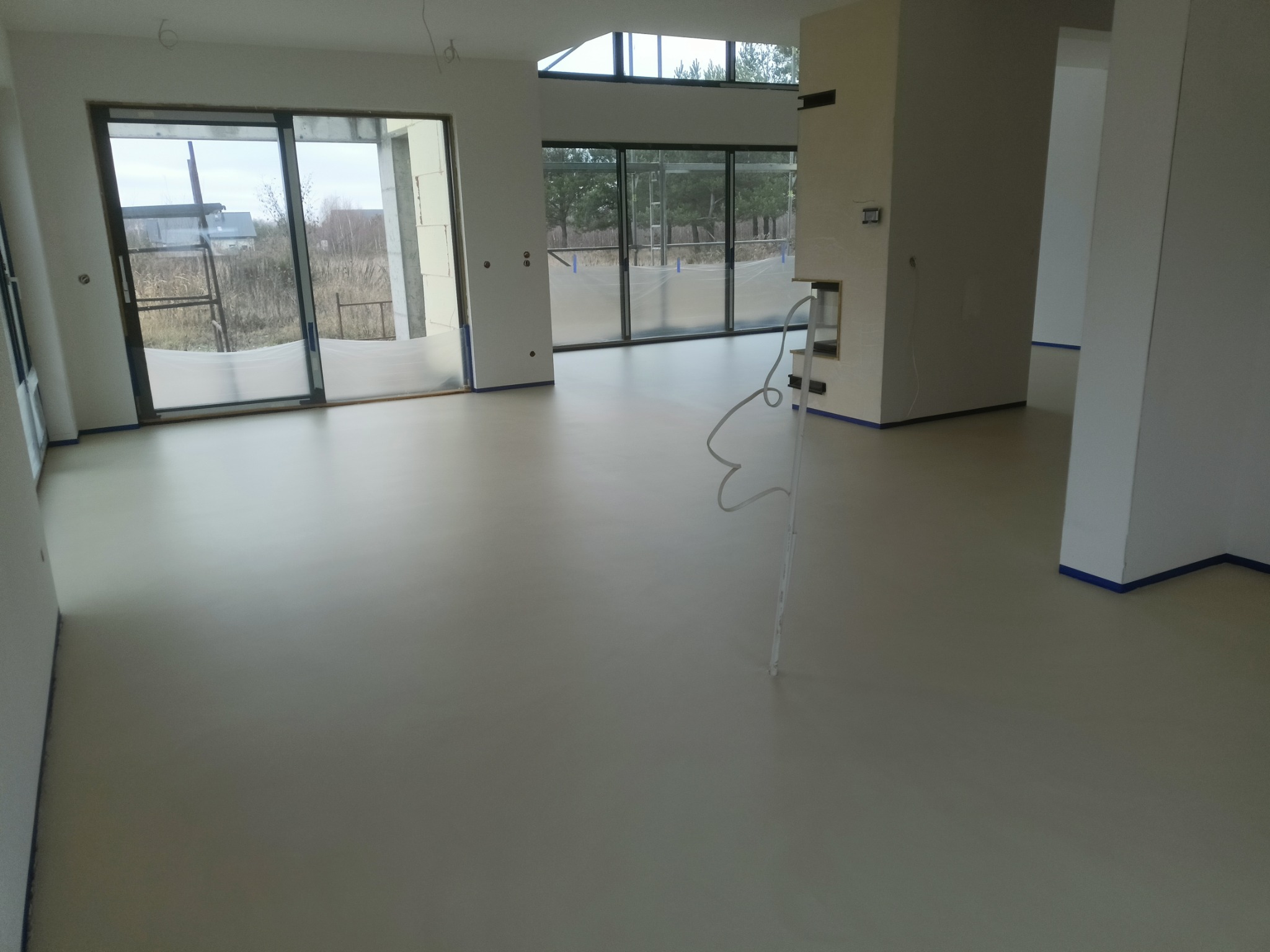 Jasna, jednolita posadzka w przestronnym, minimalistycznym wnętrzu z dużymi oknami i widokiem na zewnątrz, zabezpieczona taśmą malarską przy ścianach.