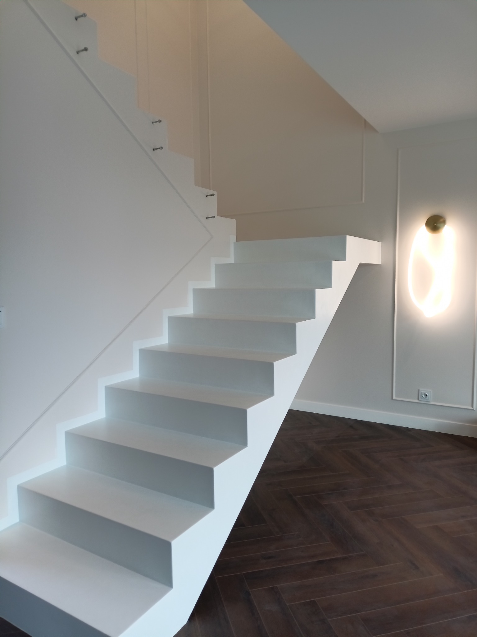 Nowoczesne, białe, wspornikowe schody z wykończeniem przypominającym beton, zamocowane do ściany za pomocą metalowych śrub, na tle parkietu ułożonego w jodełkę i dekoracyjnej lampy ściennej.