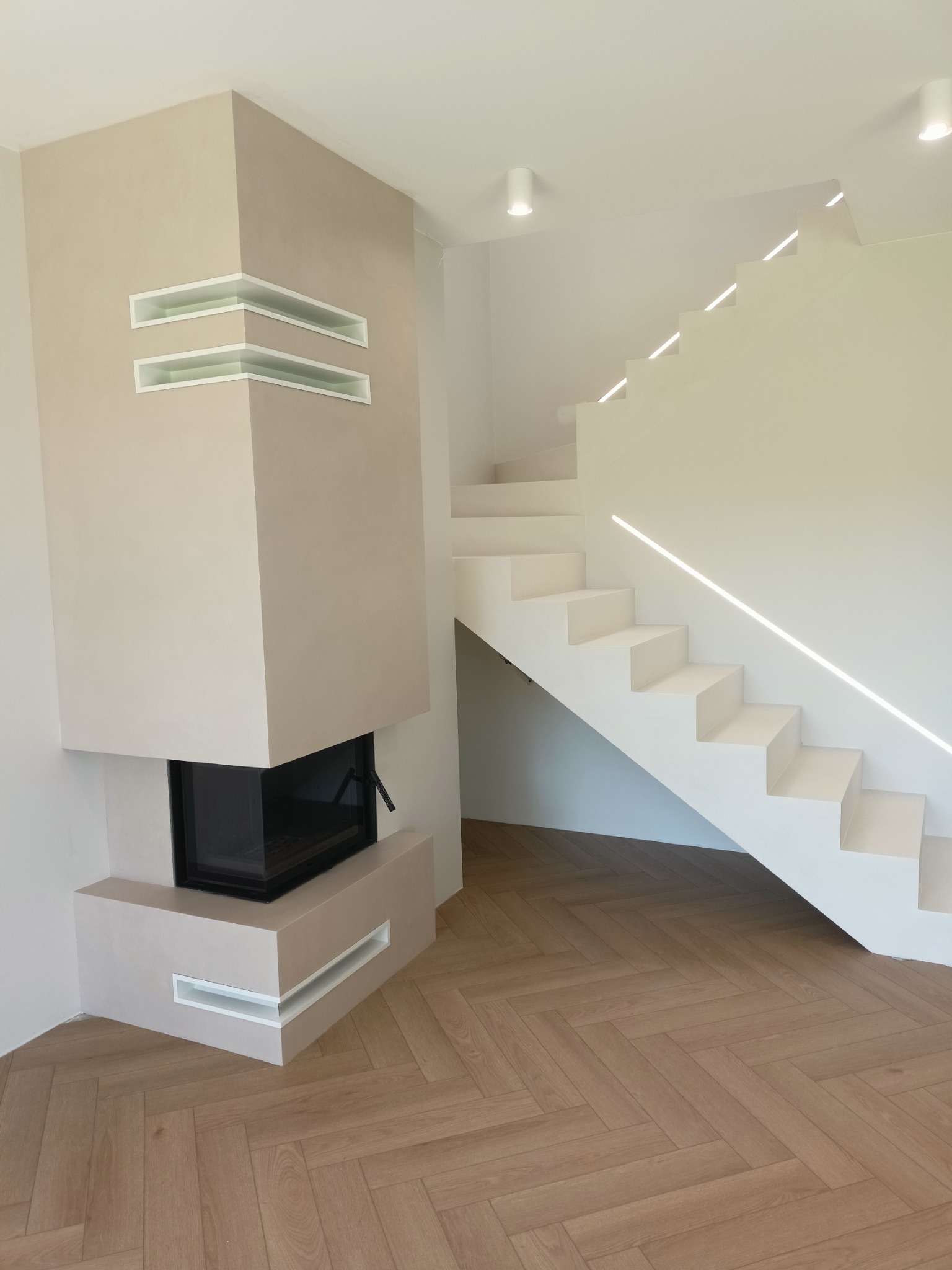 Minimalistyczne wnętrze z beżowym kominkiem z wnękami, białymi schodami z podświetleniem LED i parkietem ułożonym we wzór jodełki.