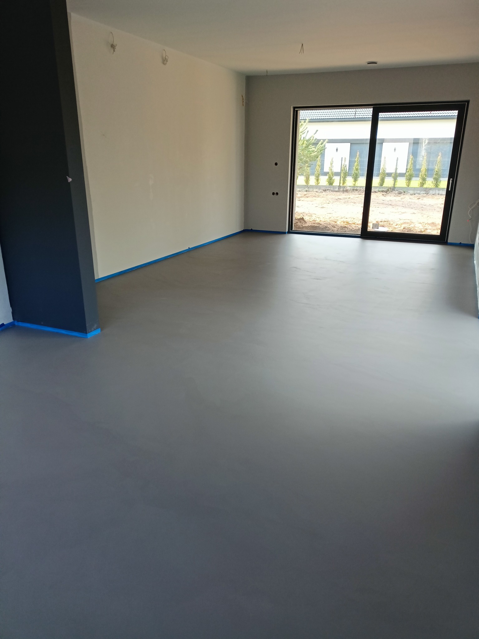 Szara, gładka podłoga w nowym domu, zabezpieczona niebieską taśmą malarską przy ścianach i filarze, duże okno przesuwne z widokiem na ogród.