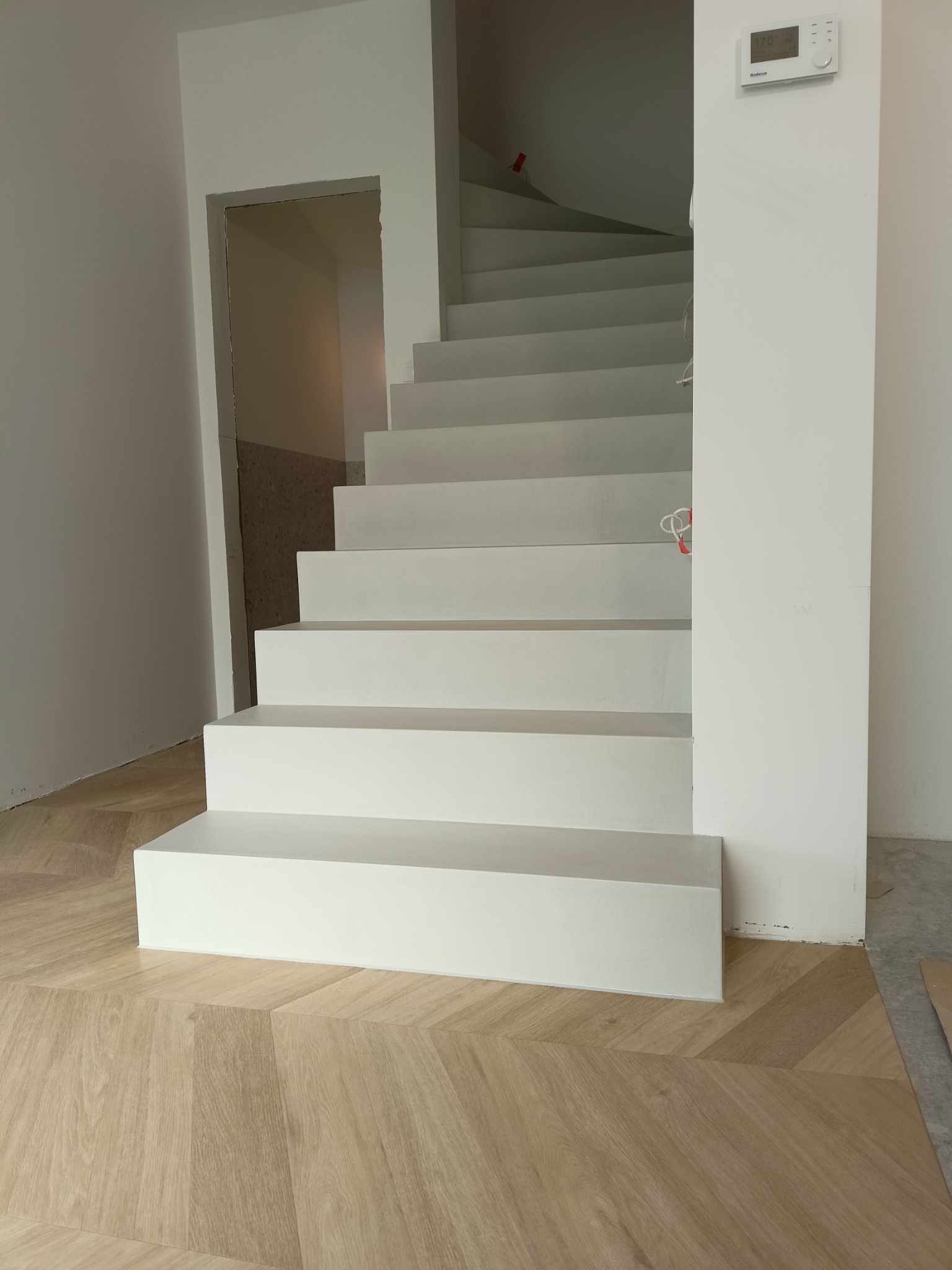 Nowoczesne, białe schody na drewnianej podłodze ułożonej we wzór jodełki, widok z dołu na pierwsze stopnie i fragment ściany z termostatem.