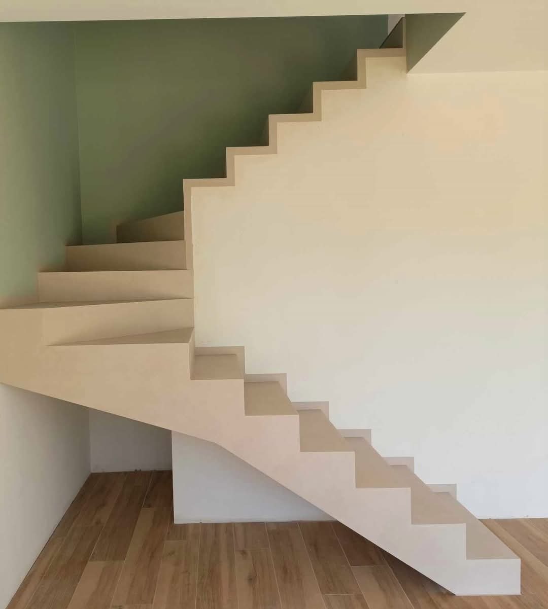 Minimalistyczne schody z beżowego betonu architektonicznego, z drewnianą podłogą i zieloną ścianą w tle. Nowoczesny design, brak poręczy.