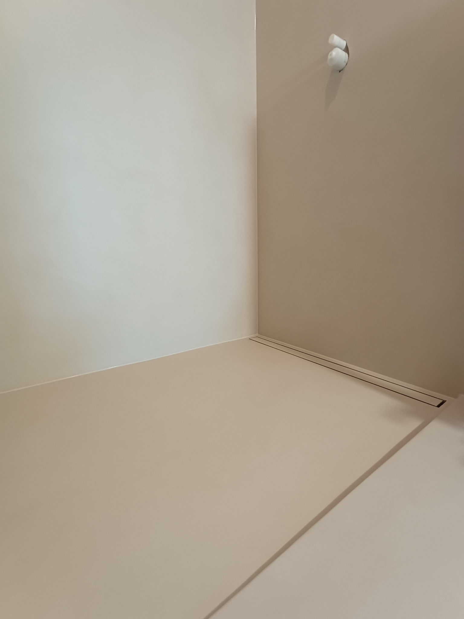 Jasna, beżowa, gładka powierzchnia w rogu pomieszczenia z odpływem liniowym. Minimalistyczne wykończenie podłogi i ścian. Widoczny fragment instalacji na ścianie.