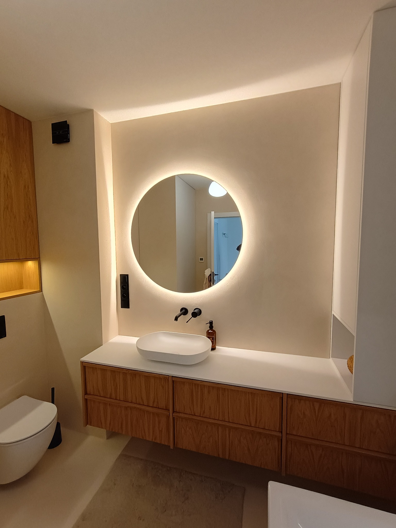 Nowoczesna łazienka z okrągłym lustrem LED, drewnianą szafką pod umywalką i beżową, jednolitą powierzchnią ścian. Minimalistyczny design.
