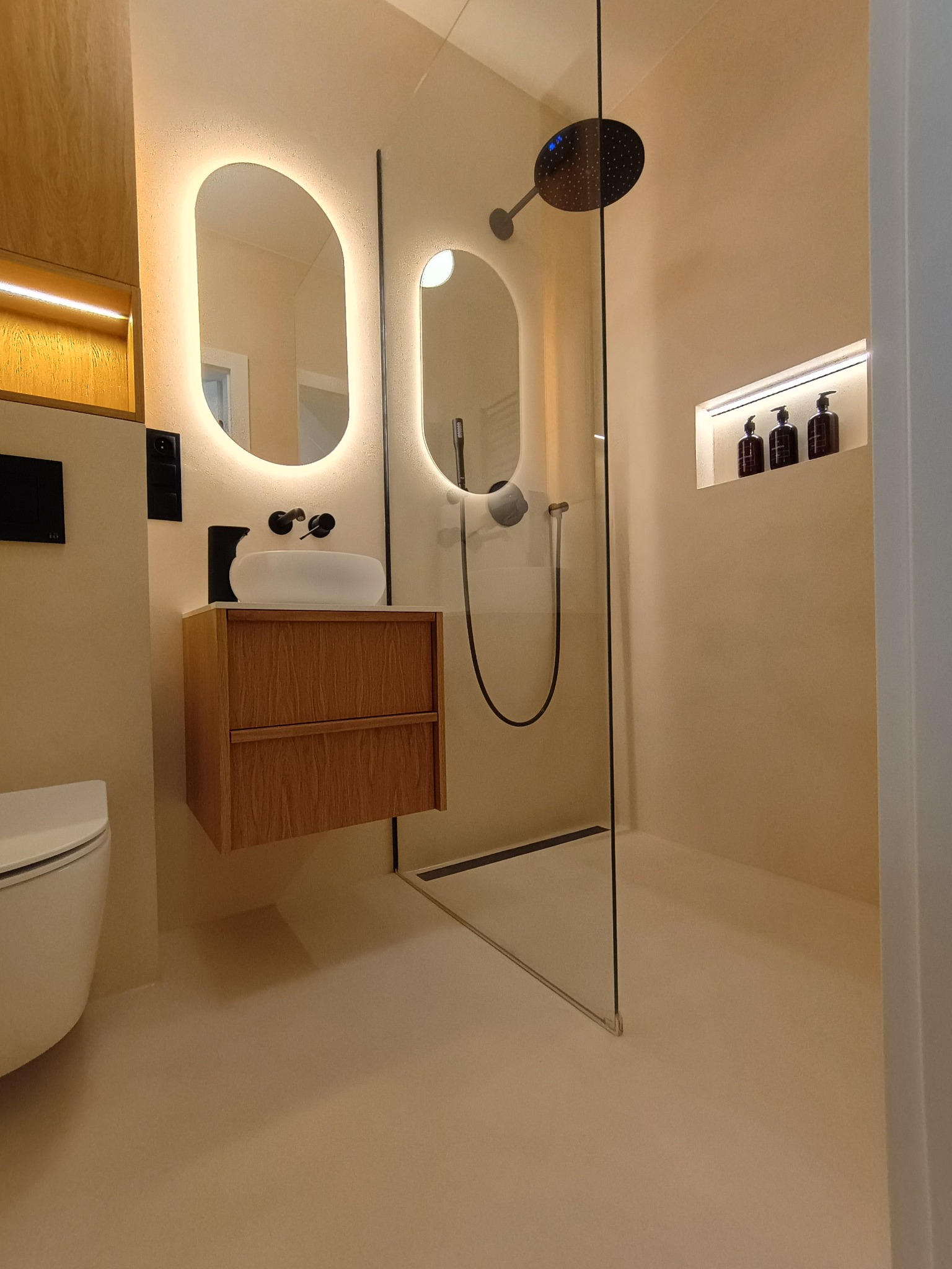 Elegancka łazienka z beżową posadzką żywiczną, kabiną prysznicową ze szklaną ścianą i oświetlonymi lustrami. Minimalistyczny design, nowoczesne wykończenie.