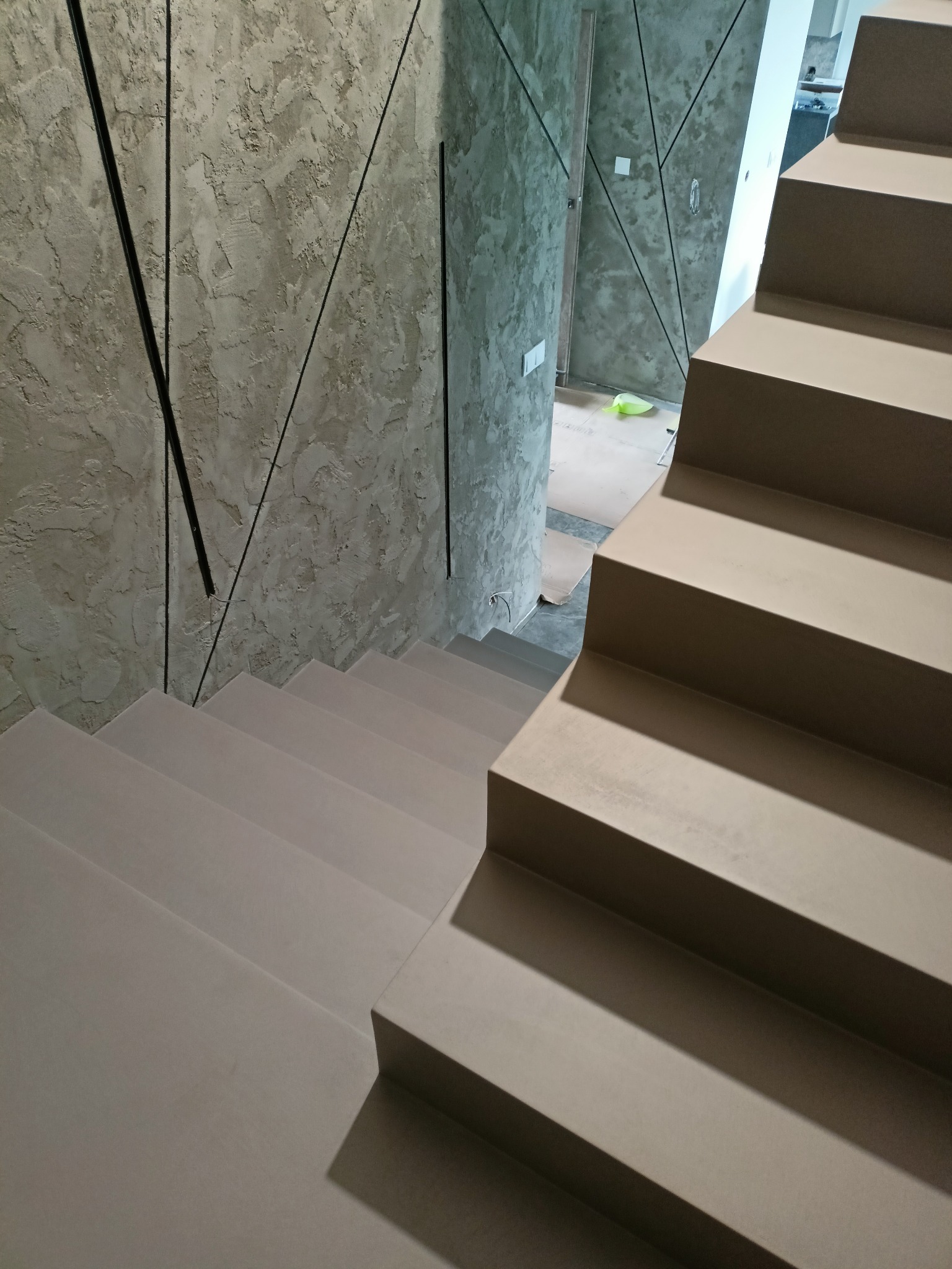 Schody wykończone jednolitą, beżową powierzchnią, z dekoracyjną ścianą w tle z geometrycznym wzorem. Widok z góry podkreśla minimalistyczny design.
