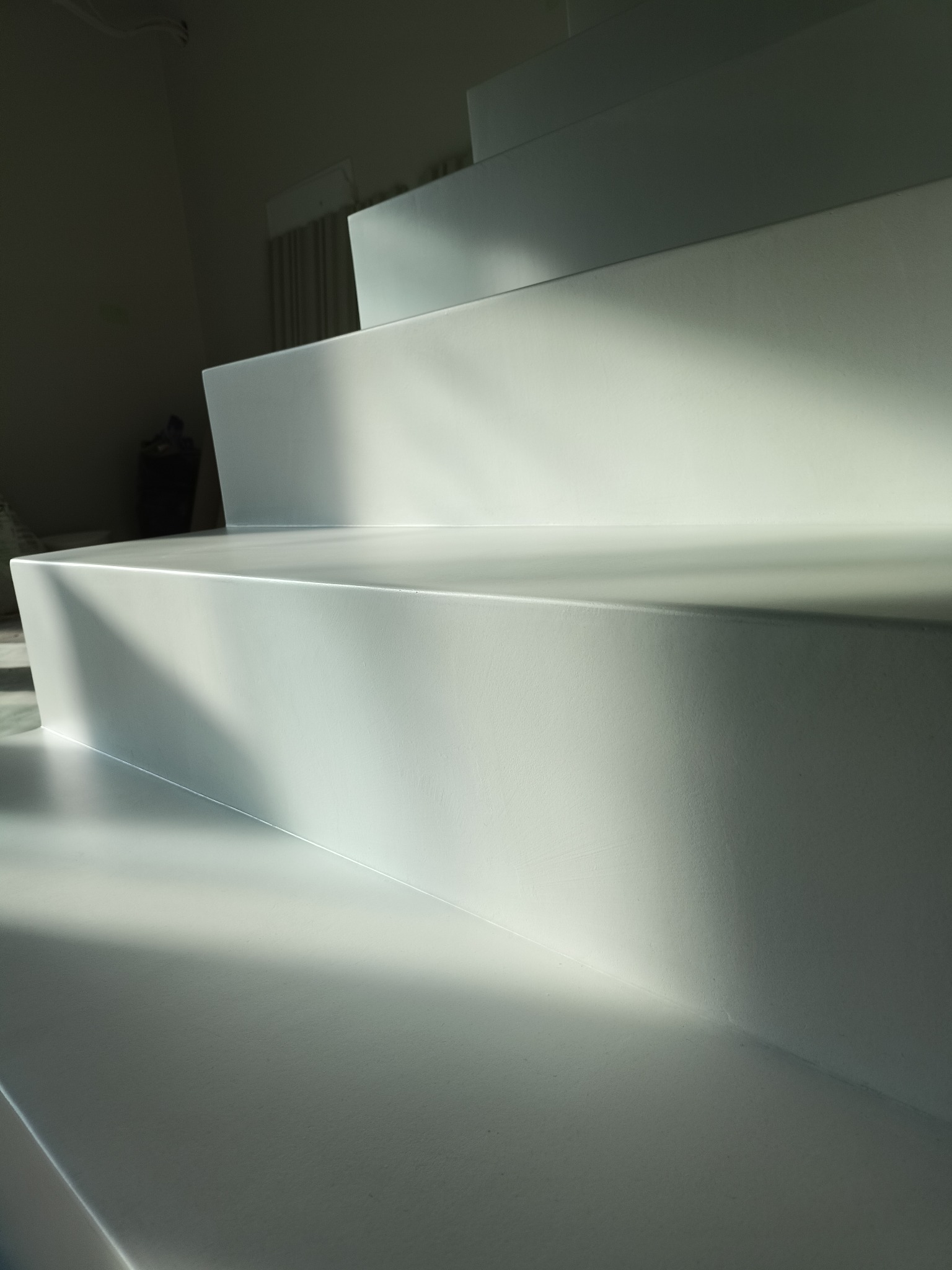 Minimalistyczne, białe schody z efektem światłocienia. Gładka powierzchnia stopni sugeruje wykończenie nowoczesnymi materiałami. W tle widoczne zasłony.