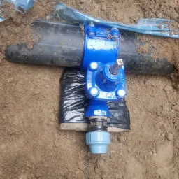 Niebieski zawór zamontowany na czarnej rurze w wykopie ziemnym, zabezpieczony folią, z widocznym fragmentem instalacji z tworzywa sztucznego.