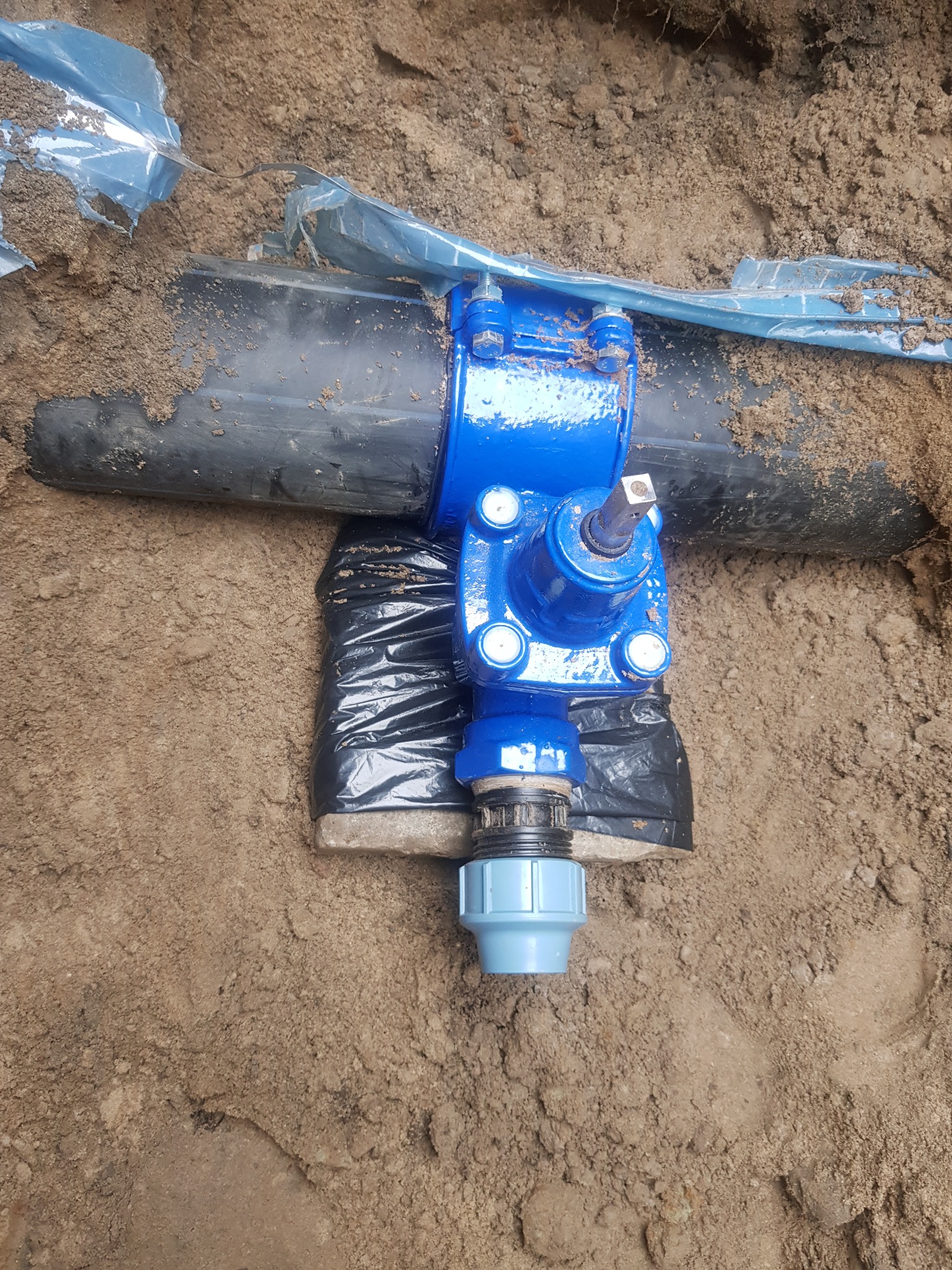 Niebieski zawór zamontowany na czarnej rurze w wykopie ziemnym, zabezpieczony folią, z widocznym fragmentem instalacji z tworzywa sztucznego.