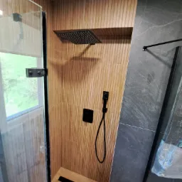 Nowoczesna kabina prysznicowa z deszczownicą i słuchawką w kolorze czarnym, wykończona płytkami imitującymi drewno i szarym marmurem.