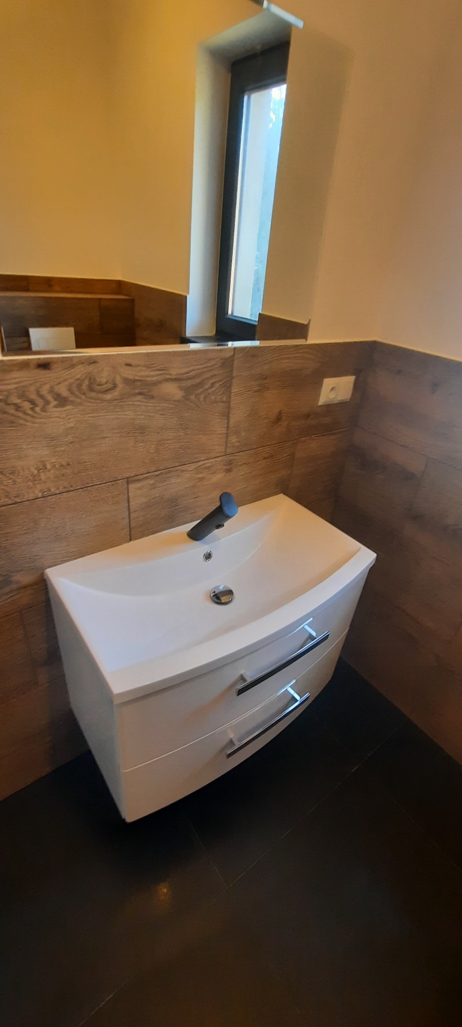Biała szafka pod umywalkę z dwiema szufladami i chromowanymi uchwytami, zamontowana w łazience z brązowymi płytkami imitującymi drewno i czarną podłogą, widoczne lustro i bateria umywalkowa.