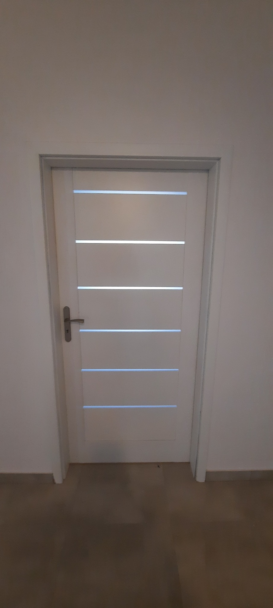 Białe drzwi wewnętrzne z poziomymi przeszkleniami, widoczne klamka i fragment podłogi.