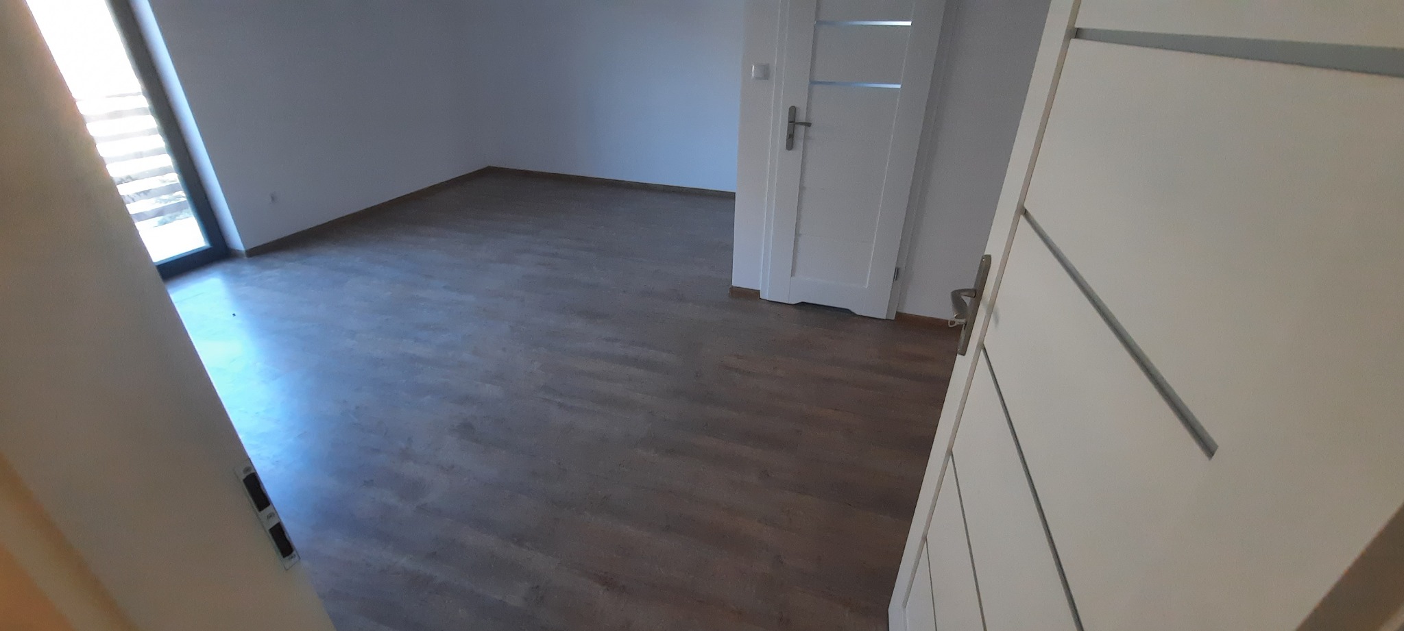 Wykończone wnętrze pokoju z panelami podłogowymi w odcieniach brązu, białe ściany i drzwi wewnętrzne z poziomymi zdobieniami, widok zza uchylonych drzwi.