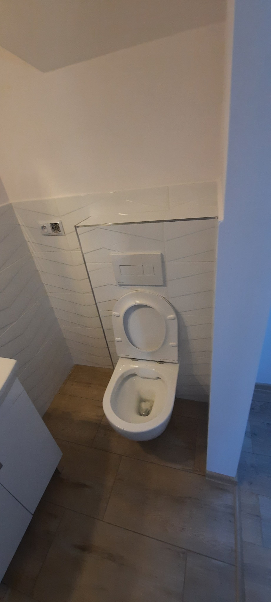 Wisząca toaleta z otwartą klapą, białe płytki z delikatnym wzorem na ścianie, drewnopodobne płytki na podłodze, widoczny fragment szafki umywalkowej i przycisk spłuczki.