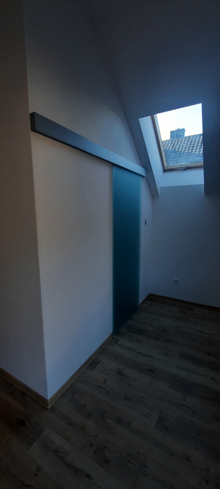 Wnętrze poddasza z drewnianą podłogą i przesuwnymi drzwiami z matowego szkła na szynie, widok na dach przez okno połaciowe.