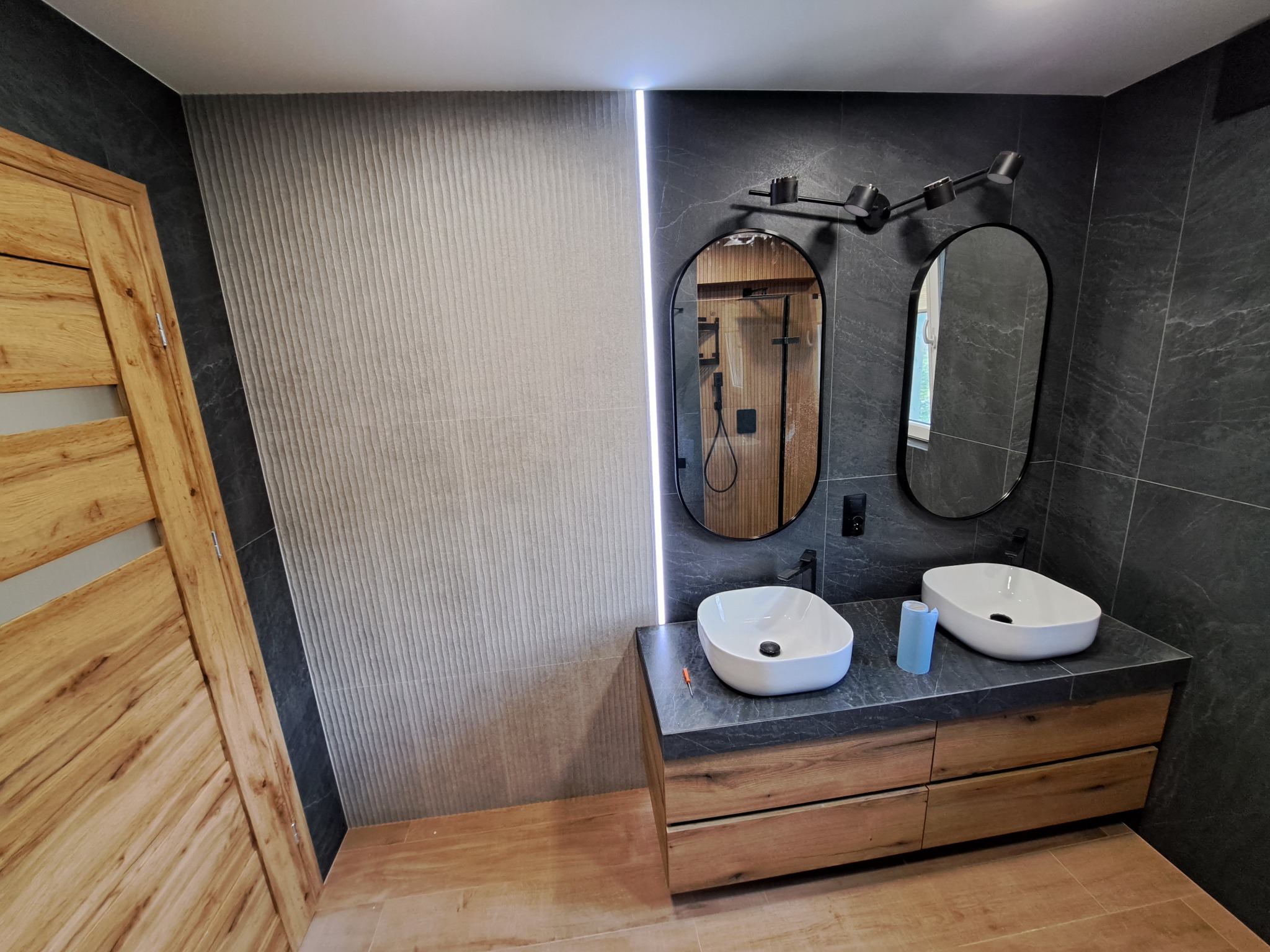 Nowoczesna łazienka z dwiema owalnymi umywalkami nablatowymi, drewnianą szafką pod umywalki z szufladami, dwoma lustrami w czarnych ramach, oświetleniem punktowym i płytkami imitującymi kamień...