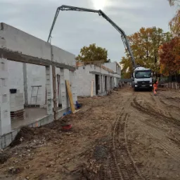 Wznoszenie ścian budynku z betonu kom&oacute;rkowego, z użyciem pompy do betonu na podwoziu ciężar&oacute;wki Mercedes, pracownik w pomarańczowym kombinezonie nadzoruje proces.
