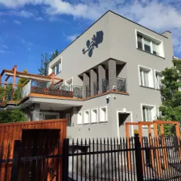 Szary dom z dekoracyjnym motywem kwiatowym na elewacji, balkonem z ozdobną balustradą i drewnianą pergolą na dachu, widok zza czarnego metalowego ogrodzenia.