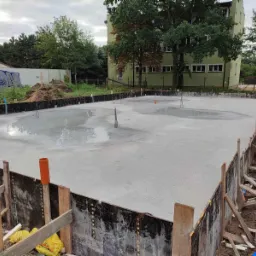 Wylany betonowy fundament z wystającymi prętami zbrojeniowymi, otoczony szalunkiem z desek i płyt, widok z góry na plac budowy w pochmurny dzień.