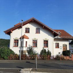 PUH MAXIM - Biały dom z czerwoną dachówką i zdobieniami roślinnymi wokół okien, otoczony pomarańczowym ogrodzeniem, widok z ulicy w słoneczny dzień.