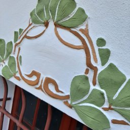 PUH MAXIM - Dekoracyjny element sztukaterii w kształcie liści i gałązek, pomalowany na zielono i brązowo, zdobiący białą ścianę nad oknem z czerwonymi kratami.