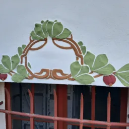 Dekoracyjny, płaskorzeźbiony ornament z motywem liści i owoców nad oknem z kratą, wykonany w odcieniach zieleni, brązu i czerwieni na białej ścianie.