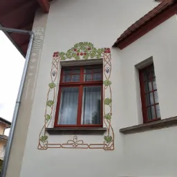 Elewacja budynku z oknami w czerwonych ramach, ozdobiona malowidłami w stylu secesyjnym z motywami roślinnymi.