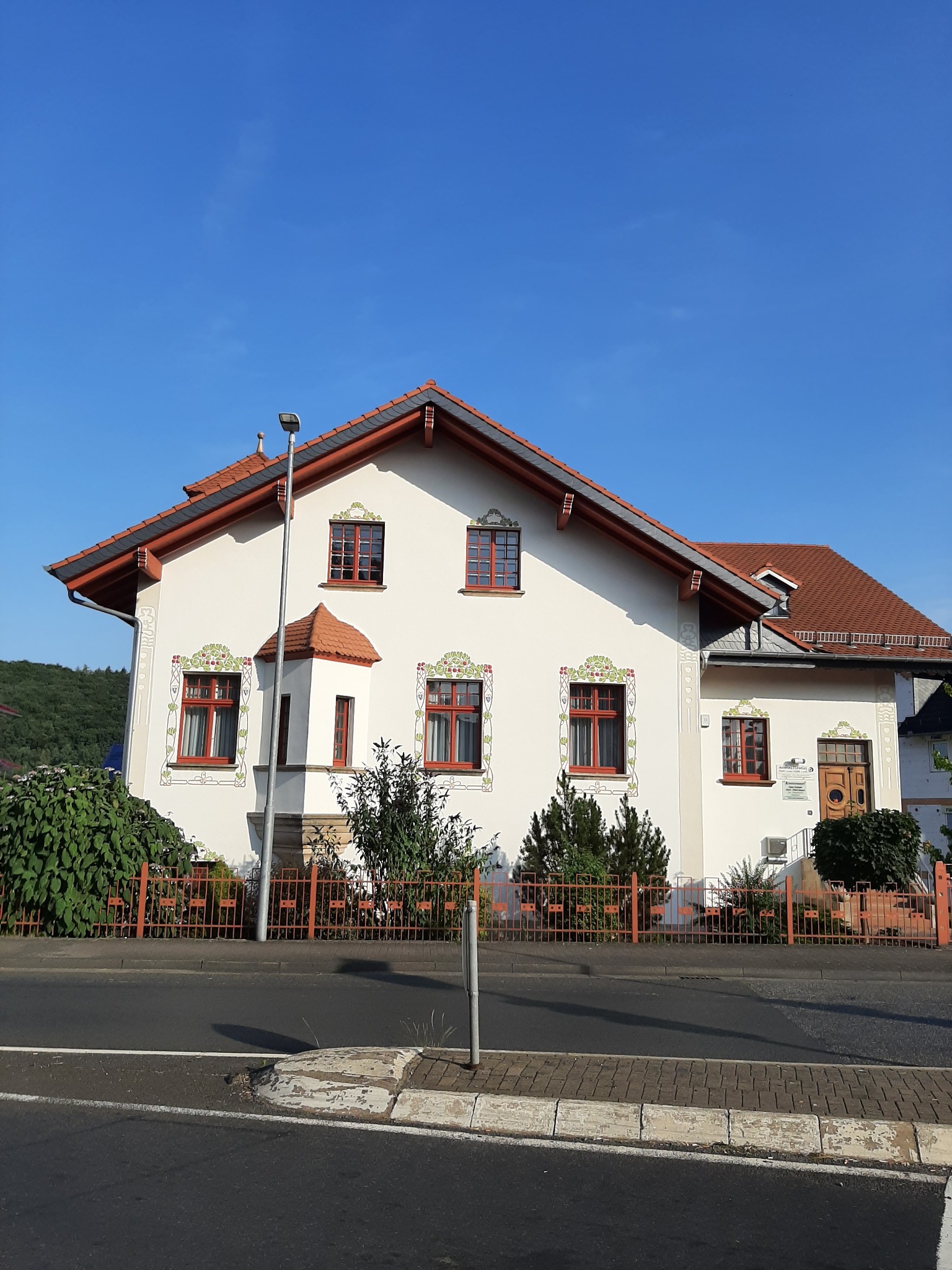 Biały dom z czerwoną dachówką i zdobieniami roślinnymi wokół okien, otoczony pomarańczowym ogrodzeniem, widok z ulicy w słoneczny dzień.