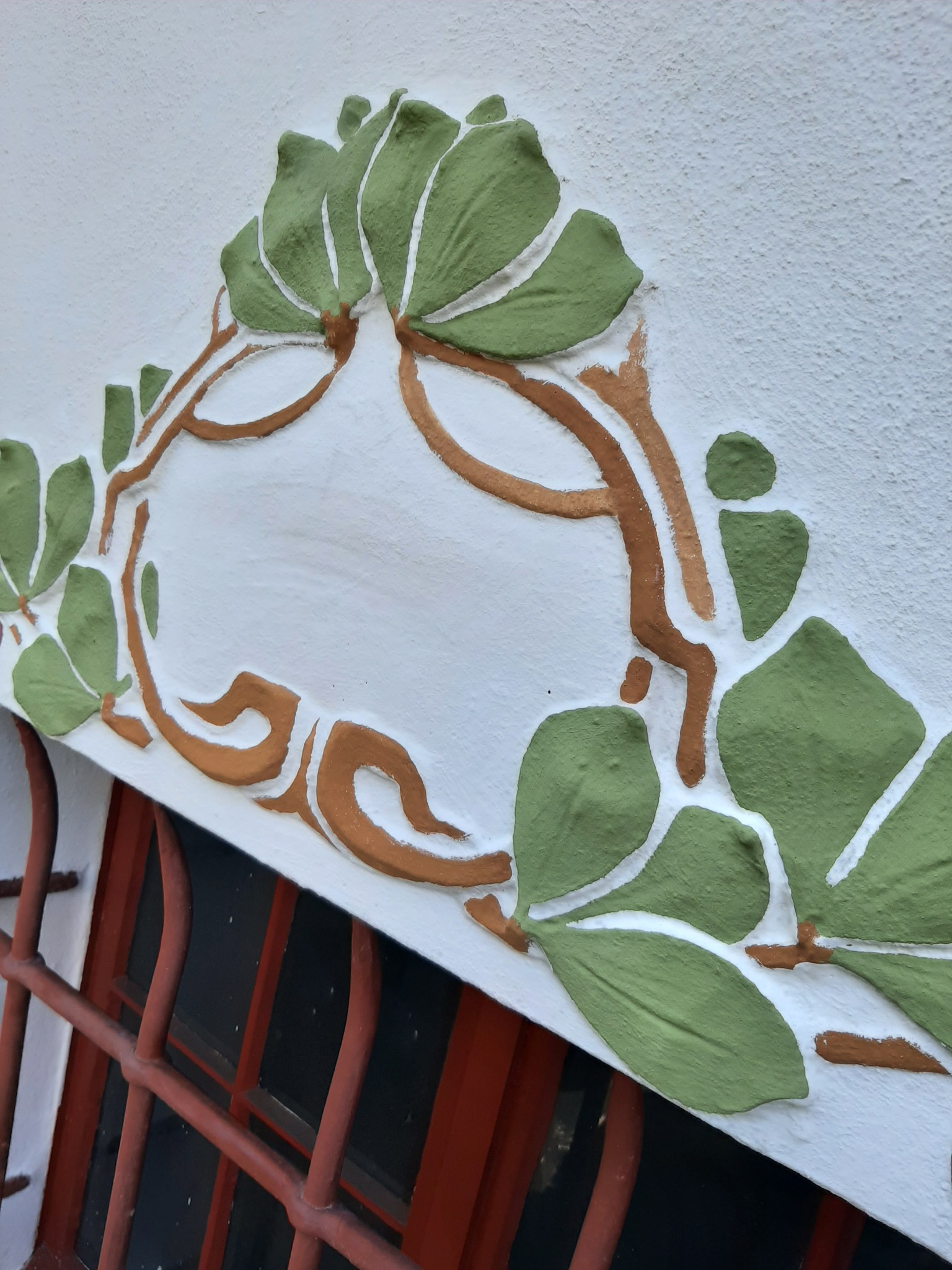 Dekoracyjny element sztukaterii w kształcie liści i gałązek, pomalowany na zielono i brązowo, zdobiący białą ścianę nad oknem z czerwonymi kratami.