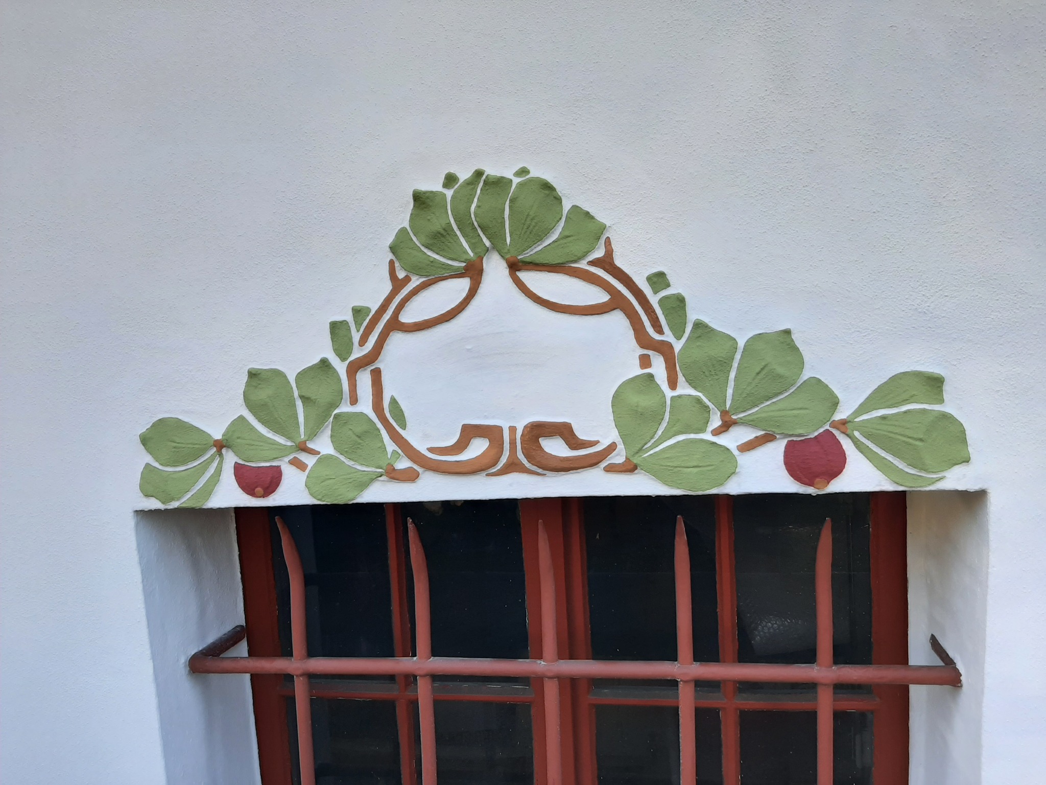 Ozdobny relief roślinny z liśćmi i owocami w kolorach zieleni, brązu i czerwieni nad oknem z metalowymi kratami w kolorze czerwonym, na białej ścianie.