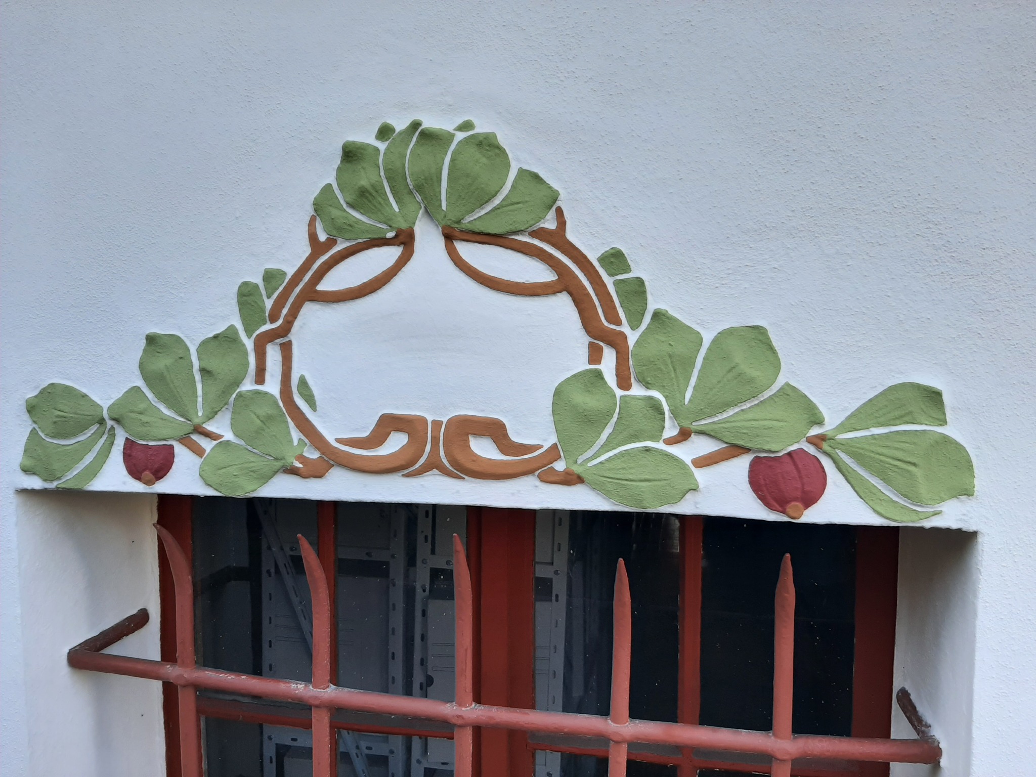 Dekoracyjny, płaskorzeźbiony ornament z motywem liści i owoców nad oknem z kratą, wykonany w odcieniach zieleni, brązu i czerwieni na białej ścianie.
