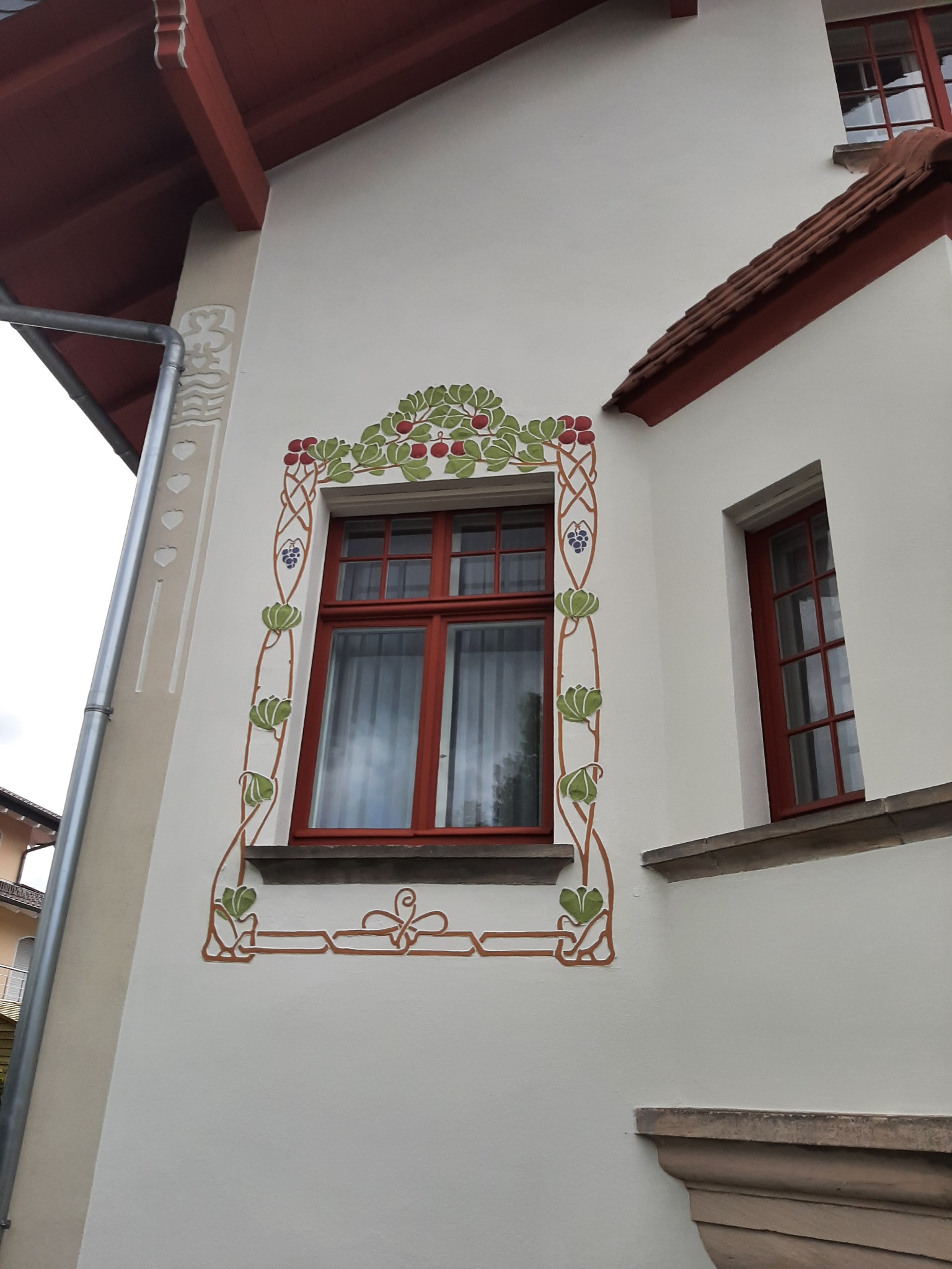 Elewacja budynku z oknami w czerwonych ramach, ozdobiona malowidłami w stylu secesyjnym z motywami roślinnymi.