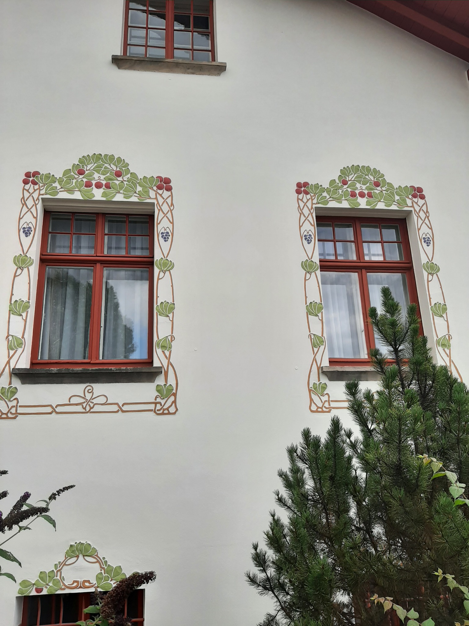 Elewacja budynku z dwoma oknami na parterze i jednym na piętrze, każde z czerwoną ramą i ozdobnymi malowidłami roślinnymi w stylu secesyjnym, otoczone zielenią krzewów.