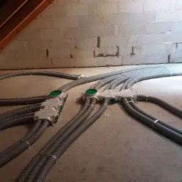 Szare, karbowane rury wentylacyjne rozchodzące się od białych rozdzielaczy z zielonymi pokrywami, ułożone na surowej podłodze w pomieszczeniu ze ścianami z białych bloczków i drewnianym skośnym...